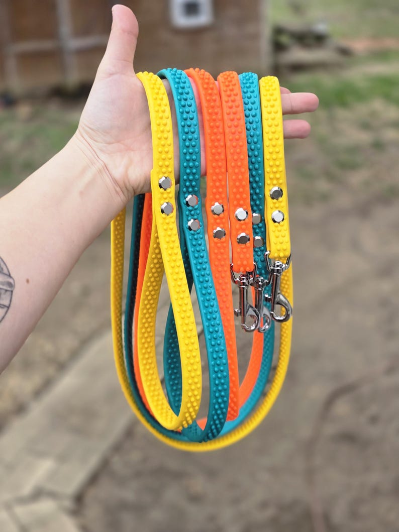 May include: A hand holds several colorful dog leashes. The leashes are yellow, teal, and orange, each with a textured surface and silver hardware. The leashes are made of a flexible material and are designed for pet owners.