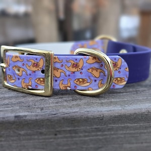 May include: A purple pet collar featuring a repeating pattern of tan pancake creatures with syrup and berries on a light purple background. The collar includes a gold-toned metal buckle and a matching gold-toned D-ring for leash attachment.