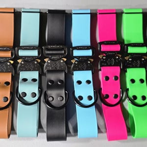 May include: A collection of colorful dog collars with black metal buckles. The collars are in various colors, including brown, teal, black, light blue, pink, and green. Each collar has a matching strap and a D-ring.