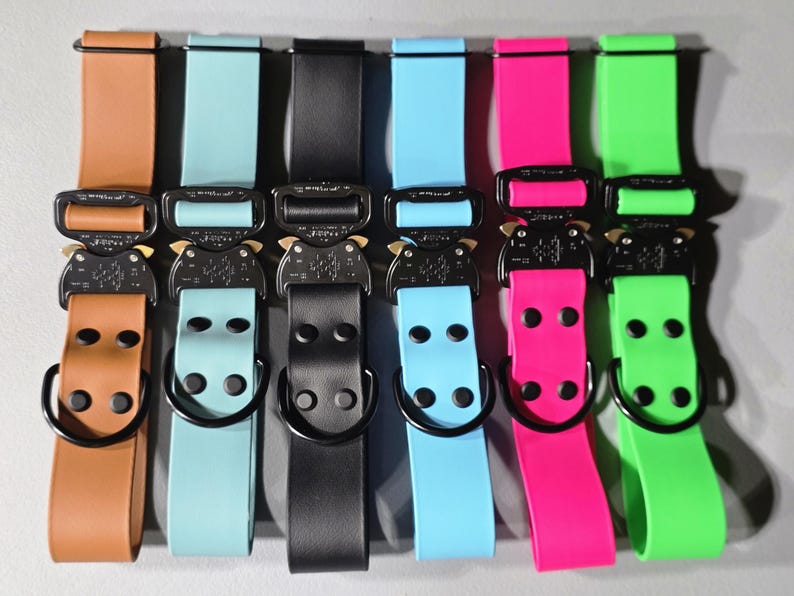 May include: A collection of colorful dog collars with black metal buckles. The collars are in various colors, including brown, teal, black, light blue, pink, and green. Each collar has a matching strap and a D-ring.