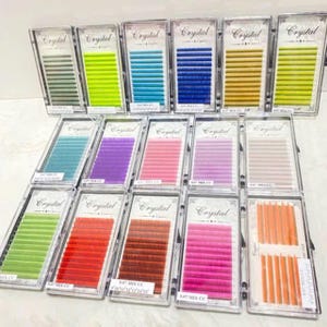 Op de afbeelding: Een verzameling van 18 individuele doosjes met kleurrijke wimperextensions. Elk doosje bevat een enkele rij wimpers in verschillende kleuren, waaronder groen, geel, blauw, paars, roze, rood, oranje en bruin. De doosjes zijn gelabeld met "Crystal" en "8-12 Mix CC".