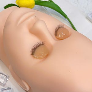 May include: A training mannequin head with applied false eyelashes. The mannequin is a light peach color, with closed eyes. The false eyelashes are black and are attached to a clear, gel-like substance. A yellow flower and green leaves are in the background.