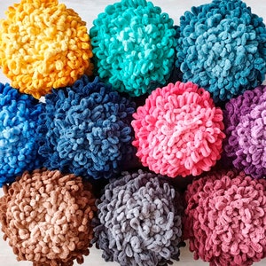 May include: A collection of eleven fluffy, round yarn balls in various colors, including yellow, teal, blue, pink, and purple. The yarn has a looped texture, creating a soft, textured appearance. The balls are arranged closely together.