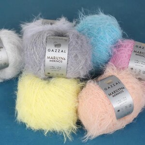 Puede incluir: Una colecci&oacute;n de ovillos de lana esponjosa en varios colores pastel, incluyendo blanco, gris, azul claro, rosa, amarillo y melocot&oacute;n. Cada ovillo tiene una etiqueta que dice "GAZZAL MARILYN & MERINOS". Ideal para tejer y hacer ganchillo.
