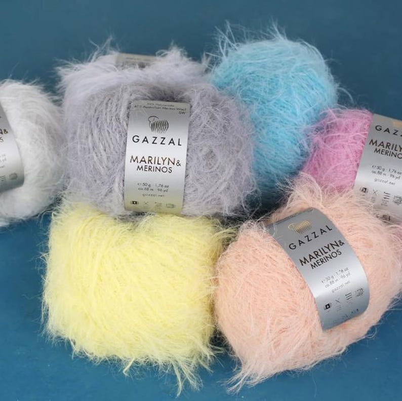 Puede incluir: Una colecci&oacute;n de ovillos de lana esponjosa en varios colores pastel, incluyendo blanco, gris, azul claro, rosa, amarillo y melocot&oacute;n. Cada ovillo tiene una etiqueta que dice "GAZZAL MARILYN & MERINOS". Ideal para tejer y hacer ganchillo.