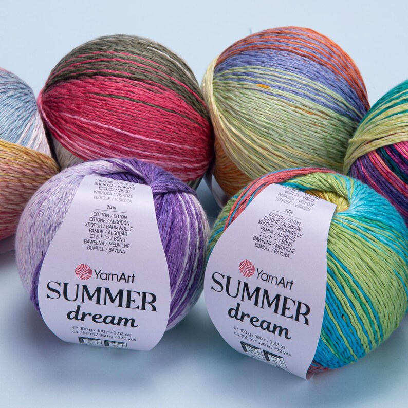 May include: Close-up of several skeins of YarnArt Summer Dream yarn. The yarn is multi-colored, with hues of pink, purple, blue, green, and yellow. Each skein has a label with the brand name and product details. The yarn is 100 grams / 3.52 oz.
