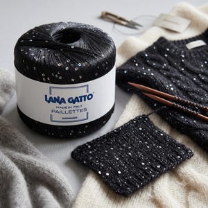 May include: A ball of black Lana Gatto Paillettes yarn with embedded silver sequins, featuring a white label with blue text. Beside it lies a knitted black swatch with sequins, a black cable-knit sweater, and two wooden knitting needles.