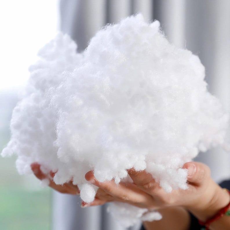 May include: A large, fluffy cloud of white polyester fiberfill. The soft, cloud-like material is held in a person's hands. This filling is often used for pillows, cushions, and stuffed animals.
