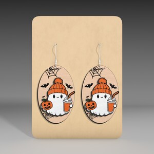 May include: A pair of Halloween-themed earrings featuring a ghost wearing a hat and holding a pumpkin and a drink. The earrings are made of wood and have a brown background with a spiderweb design.