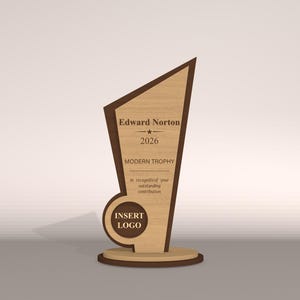 May include: A wooden trophy with a light brown finish and dark brown accents. The trophy has a unique geometric shape with the text "Edward Norton", "2026", and "MODERN TROPHY". The base includes the text "INSERT LOGO".