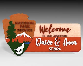 May include: A wooden sign with a red and brown background. The sign reads "Welcome to our adventure Dave & Anna ST.2024". The left side of the sign features a brown, green, and white National Park Service emblem with a wolf, a mountain, and a pine tree.