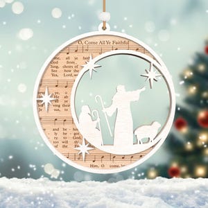 Puede incluir: Un adorno navide&ntilde;o de madera blanco con forma de media luna con una escena de la natividad en su interior. El adorno presenta un pastor, una oveja y una estrella. El adorno est&aacute; decorado con la letra del villancico "O Come All Ye Faithful".