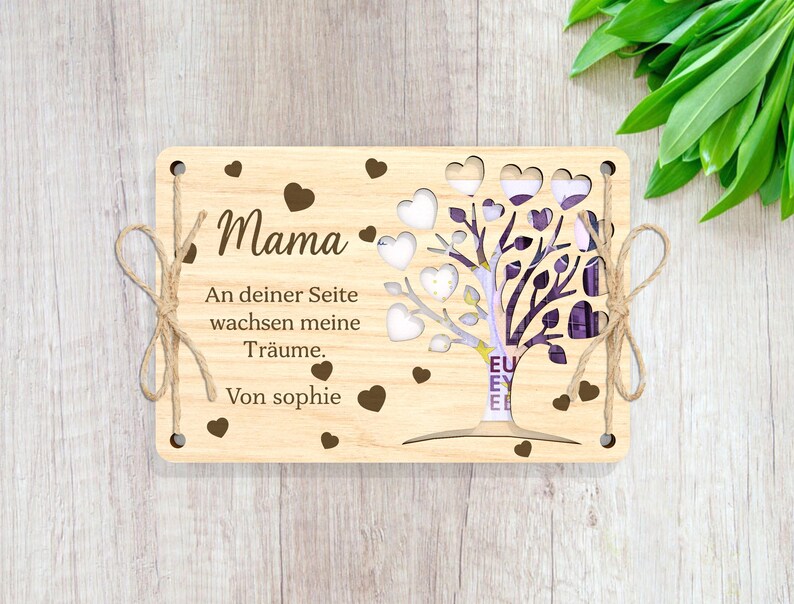 May include: A wooden card with a cutout tree design and the text "Mama. An deiner Seite wachsen meine Tr&auml;ume. Von sophie" in brown lettering. The card is tied with twine and has a heart design.