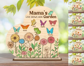 Op de afbeelding: Houten hartvormig bord met de tekst "Mama's Love Grows Here Garden". Het toont kleurrijke vlinders en bloemen, elk met een naam. Het bord staat op een houten voet, met een natuurlijke houtnerf afwerking.