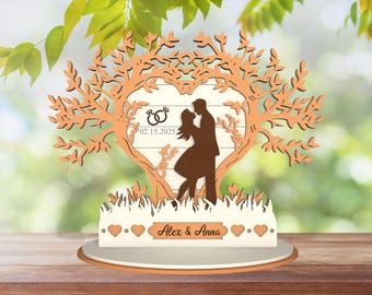 Può includere: Un topper per torta nuziale in legno con una coppia in piedi sotto un albero con un'apertura a forma di cuore. L'albero ha uno sfondo bianco con la data "02.15.2025" e due anelli. La base del topper porta i nomi "Alex & Anna" e una fila di cuori.