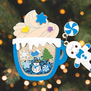 May include: A blue and white wooden ornament shaped like a mug with whipped cream and a snowman inside. The ornament has a gingerbread man hanging from it with a candy cane.