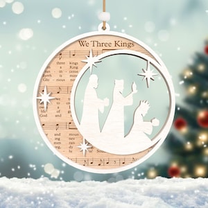 Puede incluir: Adorno navide&ntilde;o de madera blanco con un recorte de los tres reyes magos y una estrella. El adorno est&aacute; decorado con notas musicales y las palabras "We Three Kings".
