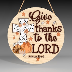 May include: A round wooden sign with a white cross and the text "Give thanks to the LORD" in brown lettering. The sign is decorated with autumnal elements such as leaves, acorns, and pumpkins. The text "PSALM 136:1" is also included.