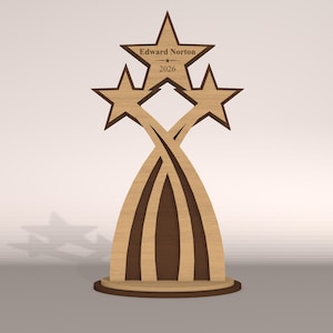 May include: A wooden trophy featuring three stars and the name "Edward Norton" with the year "2026" engraved on the top star. The design incorporates curved lines and a circular base, with a two-tone brown color scheme.