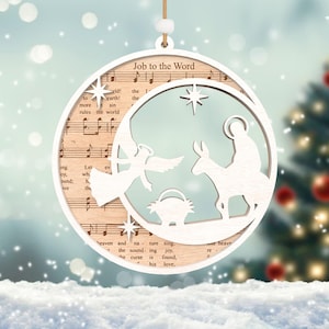 Puede incluir: Adorno navide&ntilde;o de madera blanco con un dise&ntilde;o recortado de la escena de la natividad. El adorno presenta una estrella, un &aacute;ngel, Mar&iacute;a, Jos&eacute; y un burro. El adorno est&aacute; decorado con papel de partitura musical y el texto "Job to the Word".