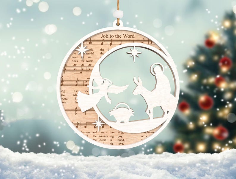 Puede incluir: Adorno navide&ntilde;o de madera blanco con un dise&ntilde;o recortado de la escena de la natividad. El adorno presenta una estrella, un &aacute;ngel, Mar&iacute;a, Jos&eacute; y un burro. El adorno est&aacute; decorado con papel de partitura musical y el texto "Job to the Word".