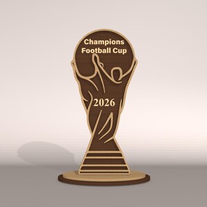 May include: A wooden Champions Football Cup trophy with the year 2026. The trophy is brown with a light brown outline and sits on a round base. The text "Champions Football Cup" is at the top.