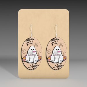 May include: A pair of wooden earrings featuring a ghost reading a book on a stack of books. The ghost is white, the books are pink and orange, and the background is wood with a spiderweb design.