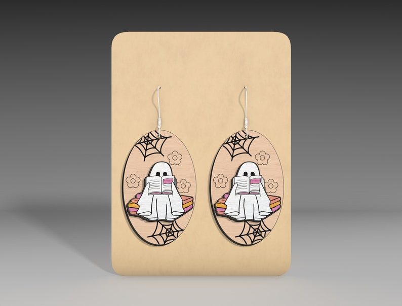 May include: A pair of wooden earrings featuring a ghost reading a book on a stack of books. The ghost is white, the books are pink and orange, and the background is wood with a spiderweb design.