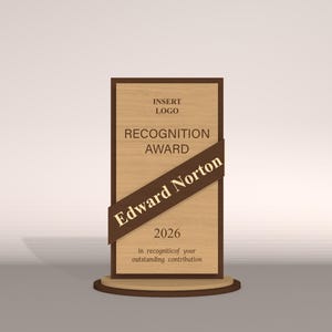 May include: A wooden recognition award with a dark brown frame and a diagonal ribbon. The text on the award reads "RECOGNITION AWARD", "Edward Norton", and "2026". The award sits on a round wooden base.