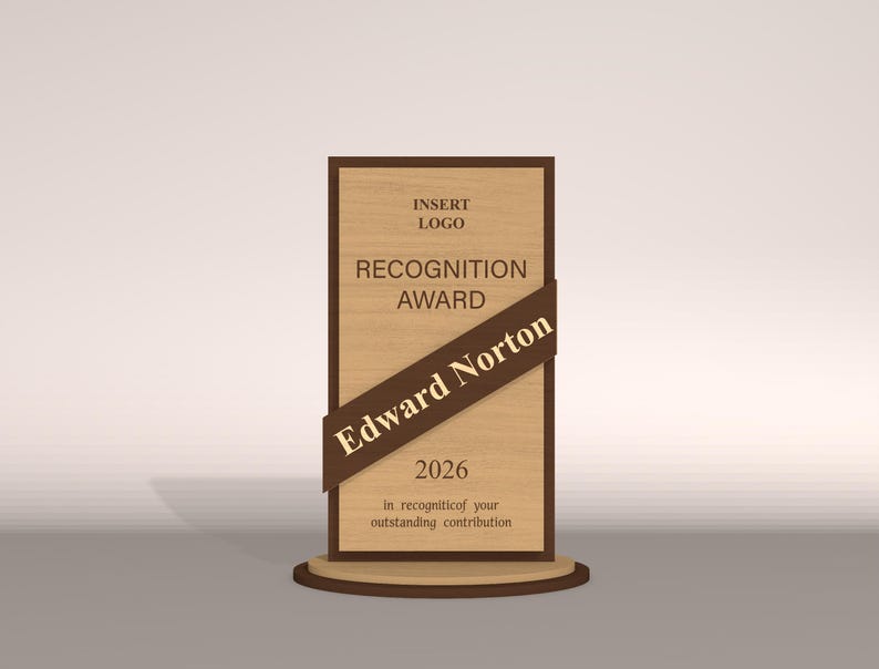May include: A wooden recognition award with a dark brown frame and a diagonal ribbon. The text on the award reads "RECOGNITION AWARD", "Edward Norton", and "2026". The award sits on a round wooden base.