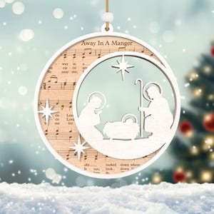 Puede incluir: Un adorno navide&ntilde;o de madera blanco con un dise&ntilde;o recortado de la escena de la natividad. El adorno est&aacute; decorado con notas musicales y las palabras "Away In A Manger".