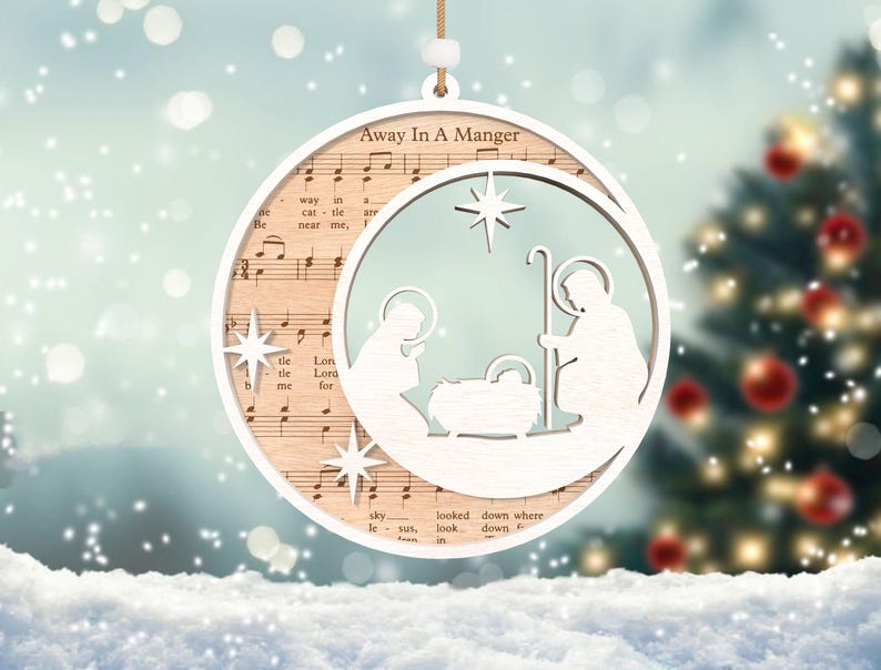 Puede incluir: Un adorno navide&ntilde;o de madera blanco con un dise&ntilde;o recortado de la escena de la natividad. El adorno est&aacute; decorado con notas musicales y las palabras "Away In A Manger".