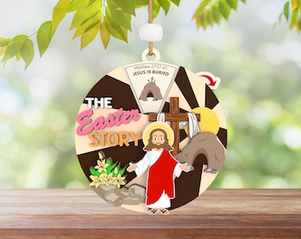 May include: A round wooden ornament with a colorful illustration of the Easter story. The ornament features a cross, a tomb, and a figure of Jesus. The text "The Easter Story" is written in pink and white letters. The text "Matthew 27:57-61 Jesus is Buried" is written in black letters.