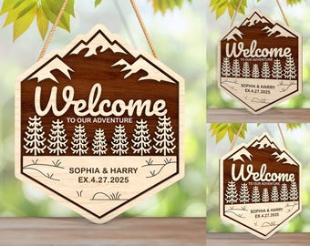 May include: A wooden welcome sign featuring a mountain range design and a forest scene. The sign reads "Welcome to our adventure" with the names "Sophia & Harry" and the date "EX. 4.27.2025".