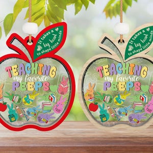 Puede incluir: Dos adornos de madera con forma de manzana, uno rojo y otro de madera natural. Ambos adornos presentan el texto "Se necesita un gran corazón para moldear las mentes pequeñas" y "Enseñando a mis pollitos favoritos" con imágenes de dibujos animados coloridos de pollitos y libros.