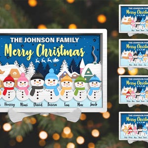 Op de afbeelding: Een witte houten kerstversiering met een blauwe achtergrond en een besneeuwde scène. De versiering toont een familie van sneeuwpoppen met verschillende gekleurde hoeden en sjaals. De tekst "THE JOHNSON FAMILY Merry Christmas" staat bovenaan de versiering.
