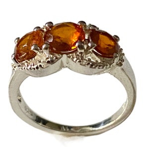 K&ouml;nnte beinhalten: Ein silberner Ring mit drei orangefarbenen Edelsteinen, die in einem dekorativen Design gefasst sind. Der Ring ist mit der Nummer 925 gekennzeichnet.