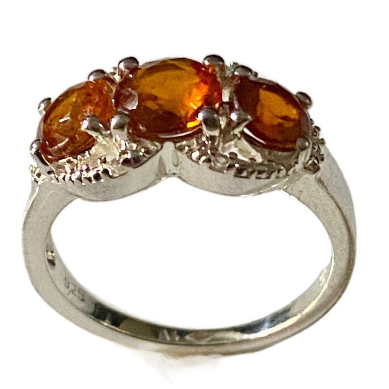 K&ouml;nnte beinhalten: Ein silberner Ring mit drei orangefarbenen Edelsteinen, die in einem dekorativen Design gefasst sind. Der Ring ist mit der Nummer 925 gekennzeichnet.
