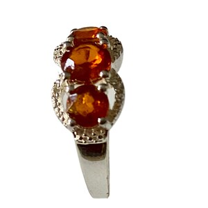 K&ouml;nnte beinhalten: Ein silberner Ring mit drei orangefarbenen Edelsteinen, die in einer Reihe angeordnet sind. Die Edelsteine sind von kleineren, klaren Steinen umgeben.