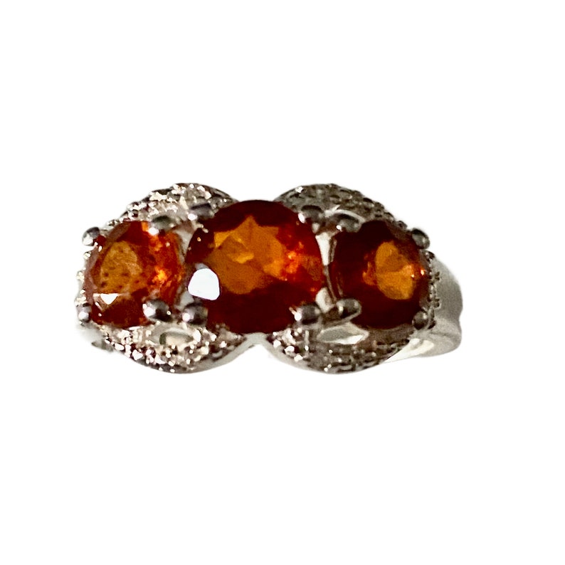 K&ouml;nnte beinhalten: Ein silberner Ring mit drei orangefarbenen Edelsteinen, die in einem dekorativen Design gefasst sind.