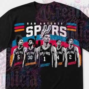 May include: Black t-shirt featuring a graphic of the San Antonio Spurs basketball team. The design includes the team name, logo, and player illustrations with their jersey numbers. The shirt has a colorful design.