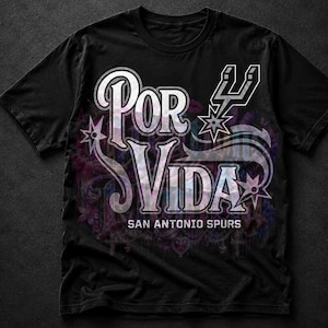 May include: Black t-shirt featuring the words "Por Vida" in a stylized font, with "San Antonio Spurs" below. The design includes a Spurs logo and decorative elements in white, purple, and blue.