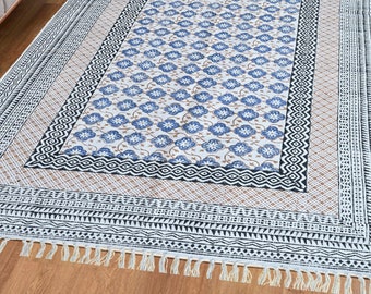 May include: A white cotton rug with a blue and brown floral pattern. The rug has a black and white geometric border and fringe on all sides.