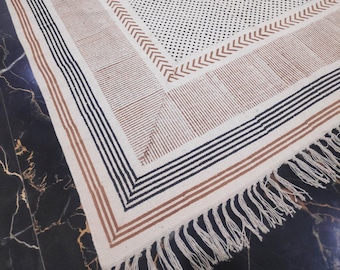 May include: A white cotton rug with a black and brown geometric pattern. The rug has a fringe border and is made of natural fibers.