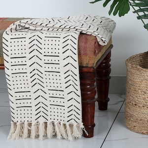 Op de afbeelding: Een witte deken met zwarte geometrische patronen en franjes. De deken is over een houten bank gelegd met een kleurrijk, gepatroneerd kussen.