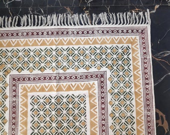May include: A white cotton rug with a geometric pattern in shades of brown, green, and yellow. The rug has a fringe border.