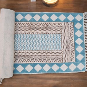 Puede incluir: Una alfombra rectangular parcialmente enrollada con un borde azul con formas de diamantes blancos. El centro de la alfombra tiene un patrón geométrico en marrón y azul, con una franja blanca en el borde derecho. La alfombra está sobre una superficie de madera.