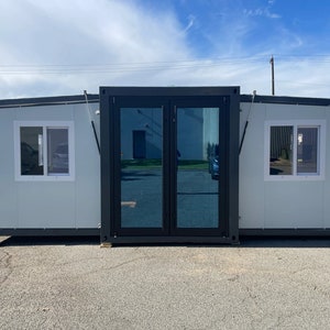 May include: A gray modular building with two windows and a large glass door. The building is made of metal panels and has a black trim.