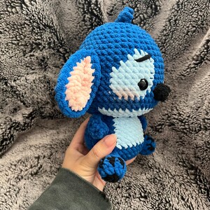 May include: Blue and pink crocheted Stitch plush toy with black eyes and a black nose. The toy is sitting on a fuzzy gray surface.