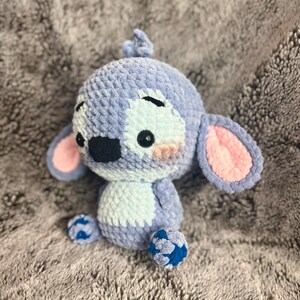 May include: A blue and white crocheted plush toy with pink ears and blue feet. The toy has a stitched-on face with black eyes and a black nose.